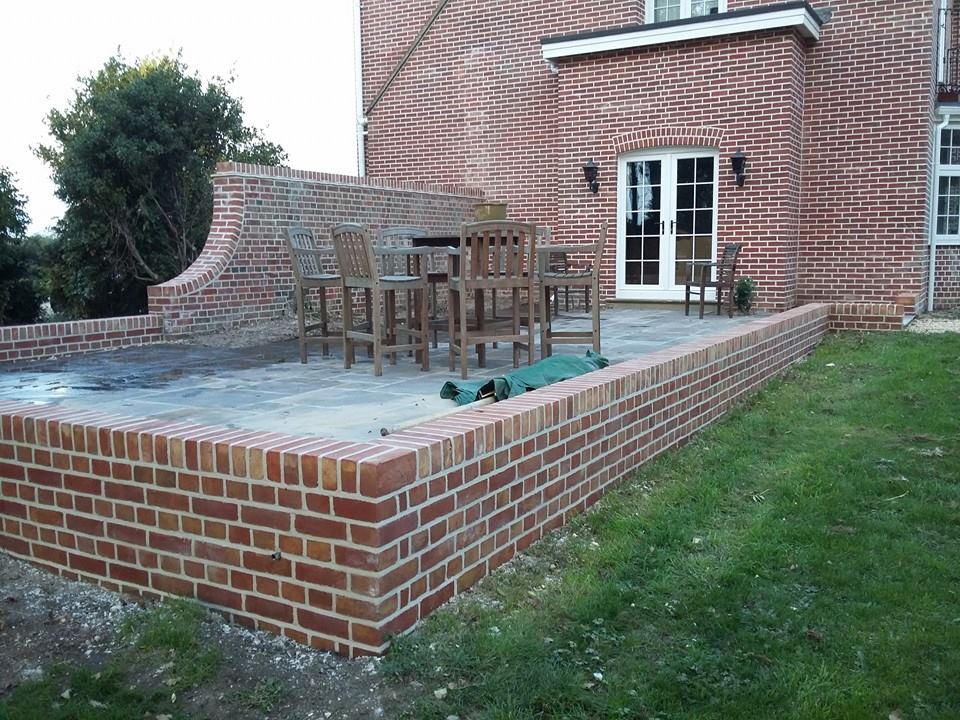 A red brick retaining wall with a brick-on-edge coping, enclosing a raised paved terrace with garden furniture